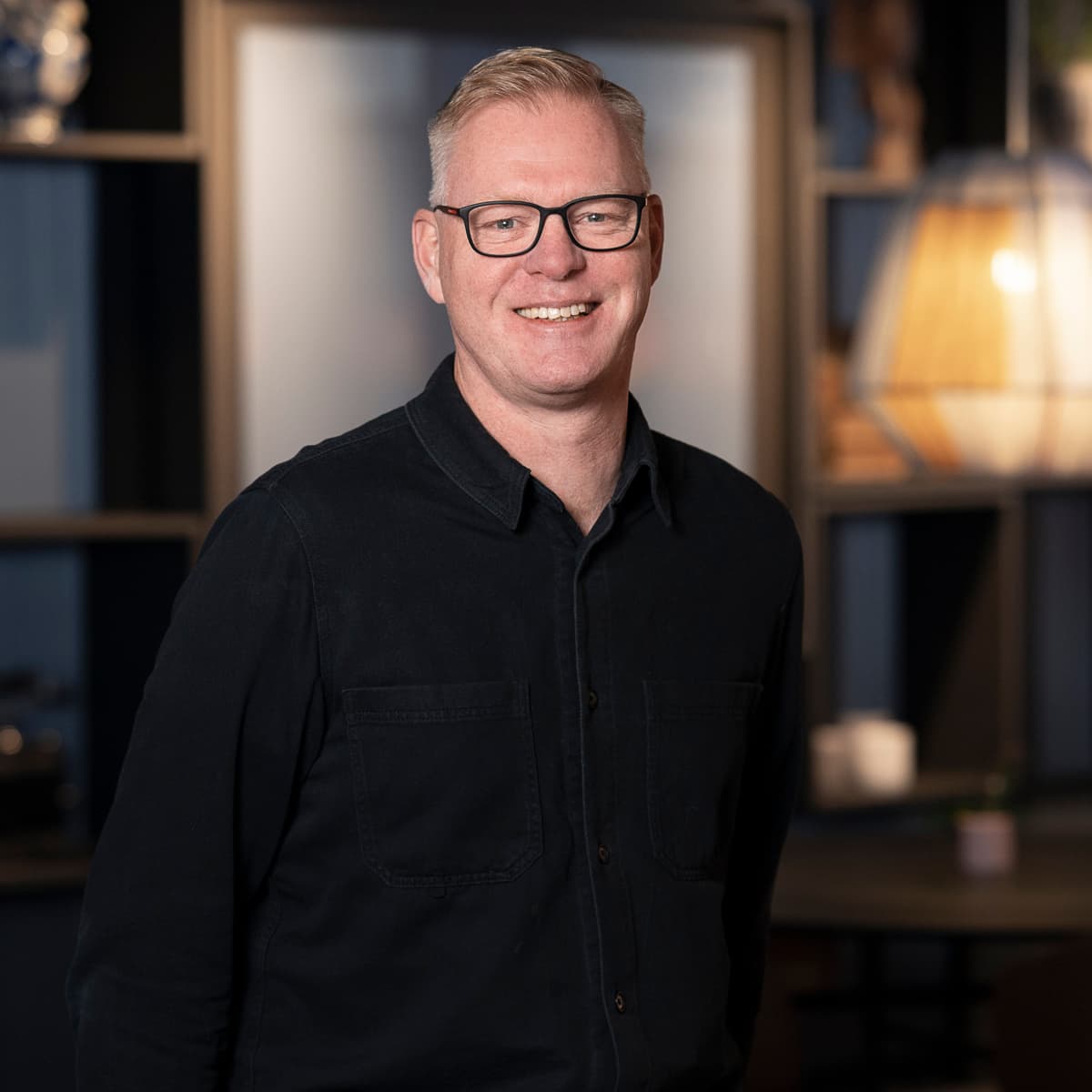 En man i svart skjorta och glasögon står i ett rum.