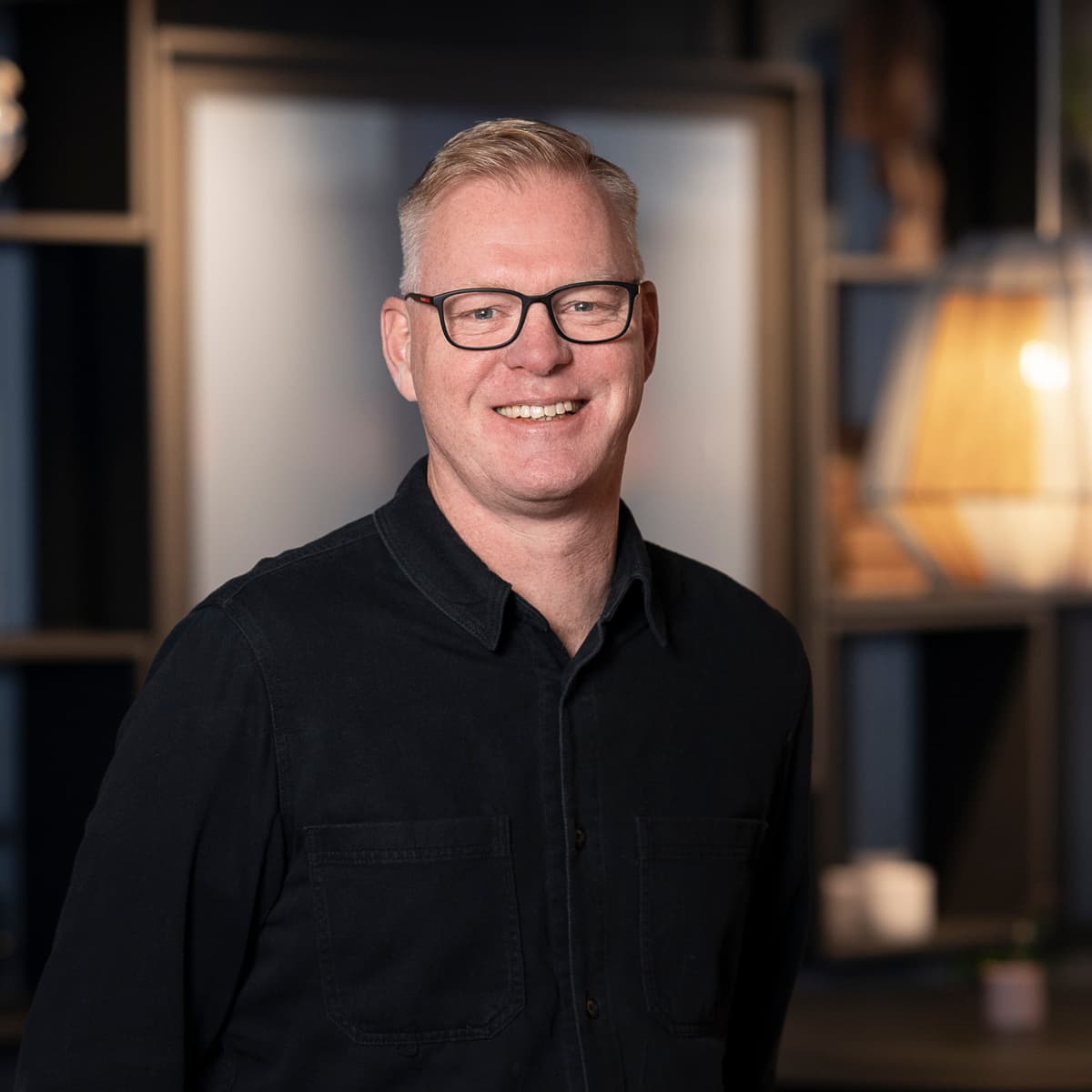 En man i svart skjorta och glasögon står i ett rum.