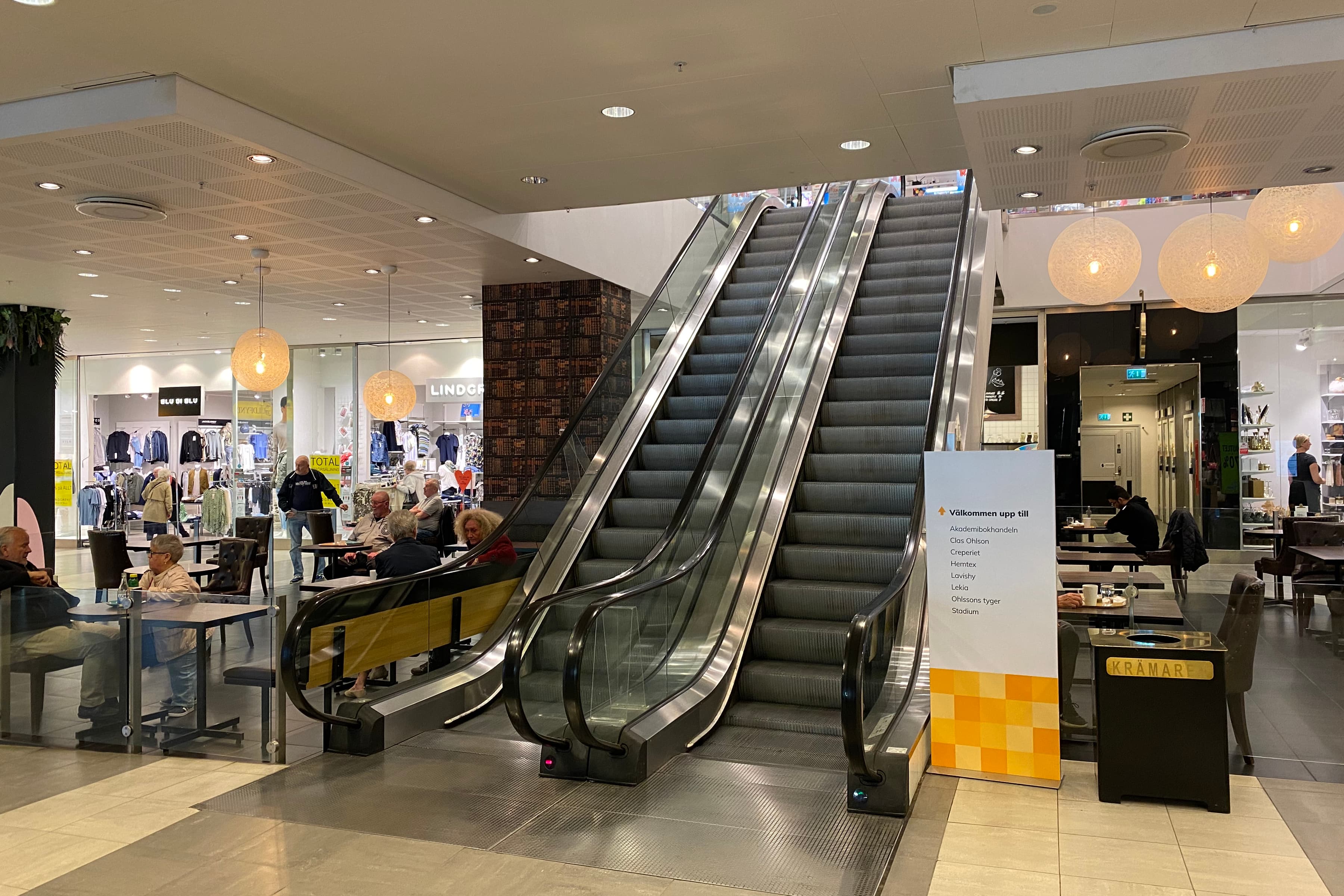 En rulltrappa i ett köpcentrum med människor som sitter vid bord.